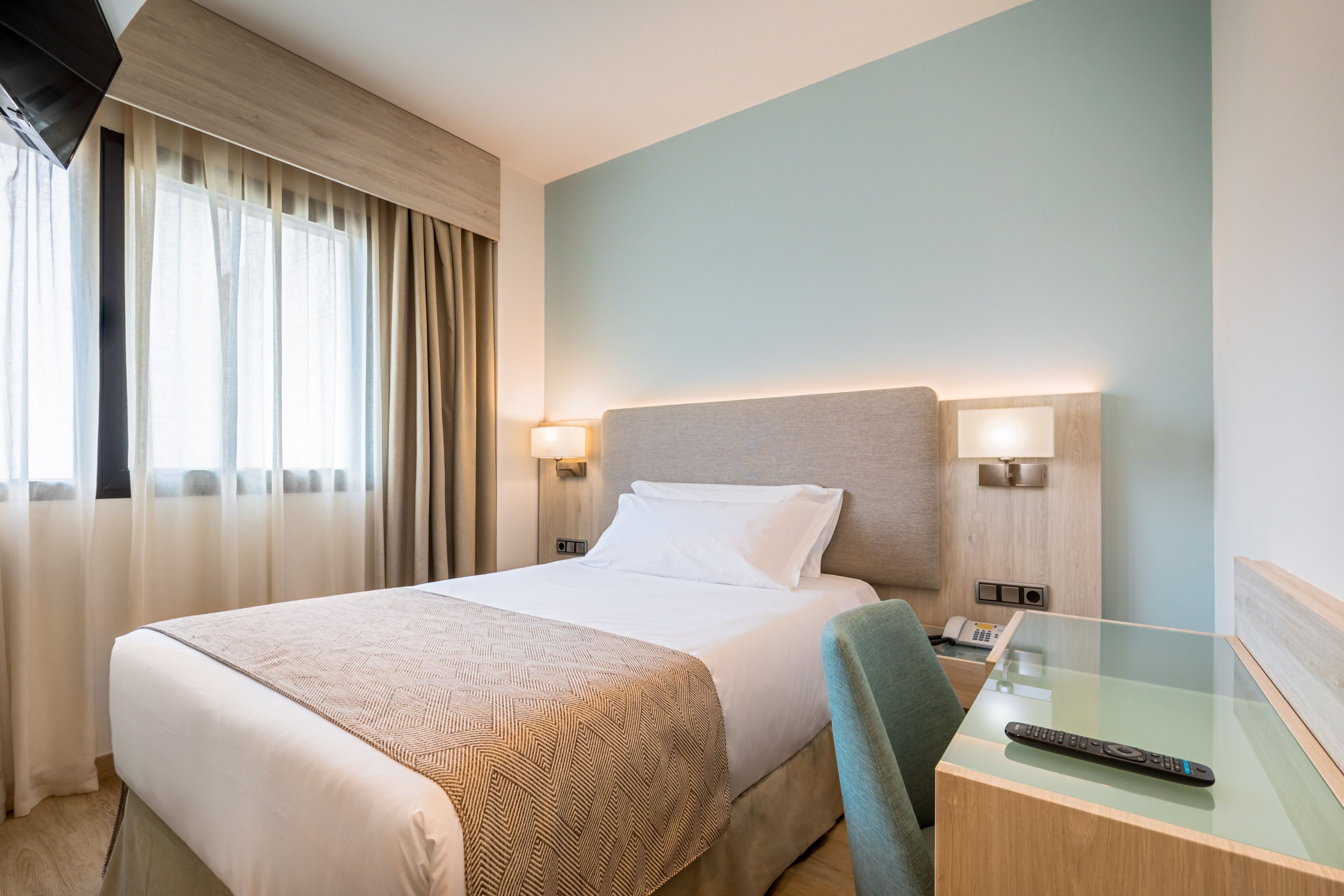 Een hotelkamer met een bed, bedlampjes, een bureau met een afstandsbediening en een raam met gordijnen.