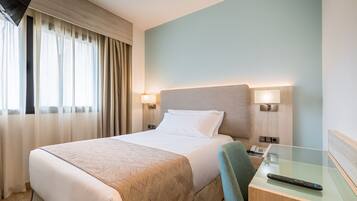 Een hotelkamer met een bed, bedlampjes, een bureau met een afstandsbediening en een raam met gordijnen.