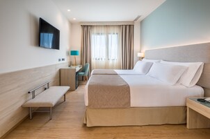 Una habitación de hotel con una cama, una banca, un escritorio, un televisor y una ventana con cortinas.