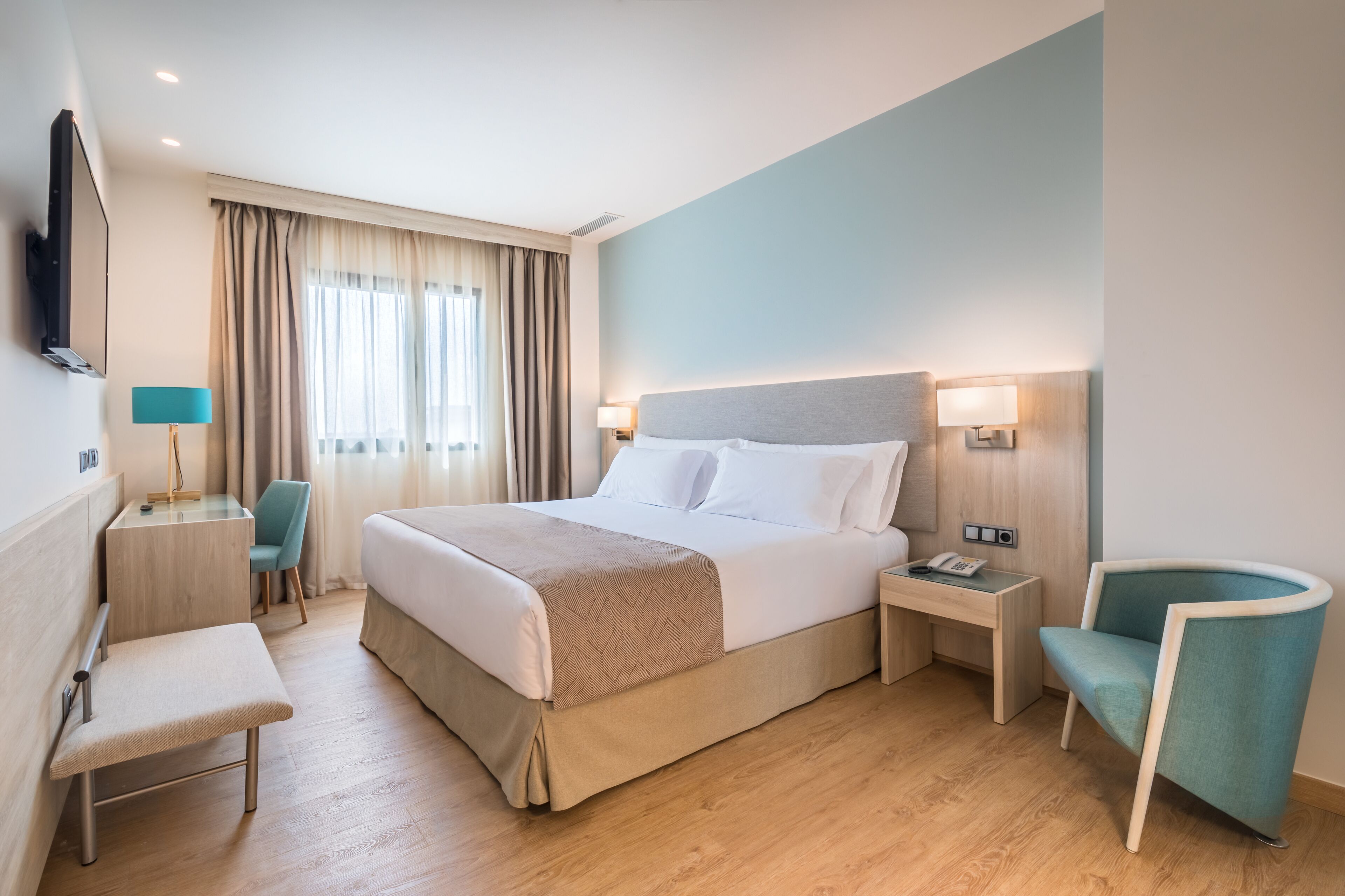 Una habitació d'hotel amb un llit, taules de nit, un escriptori, una cadira i un televisor penjat a la paret.
