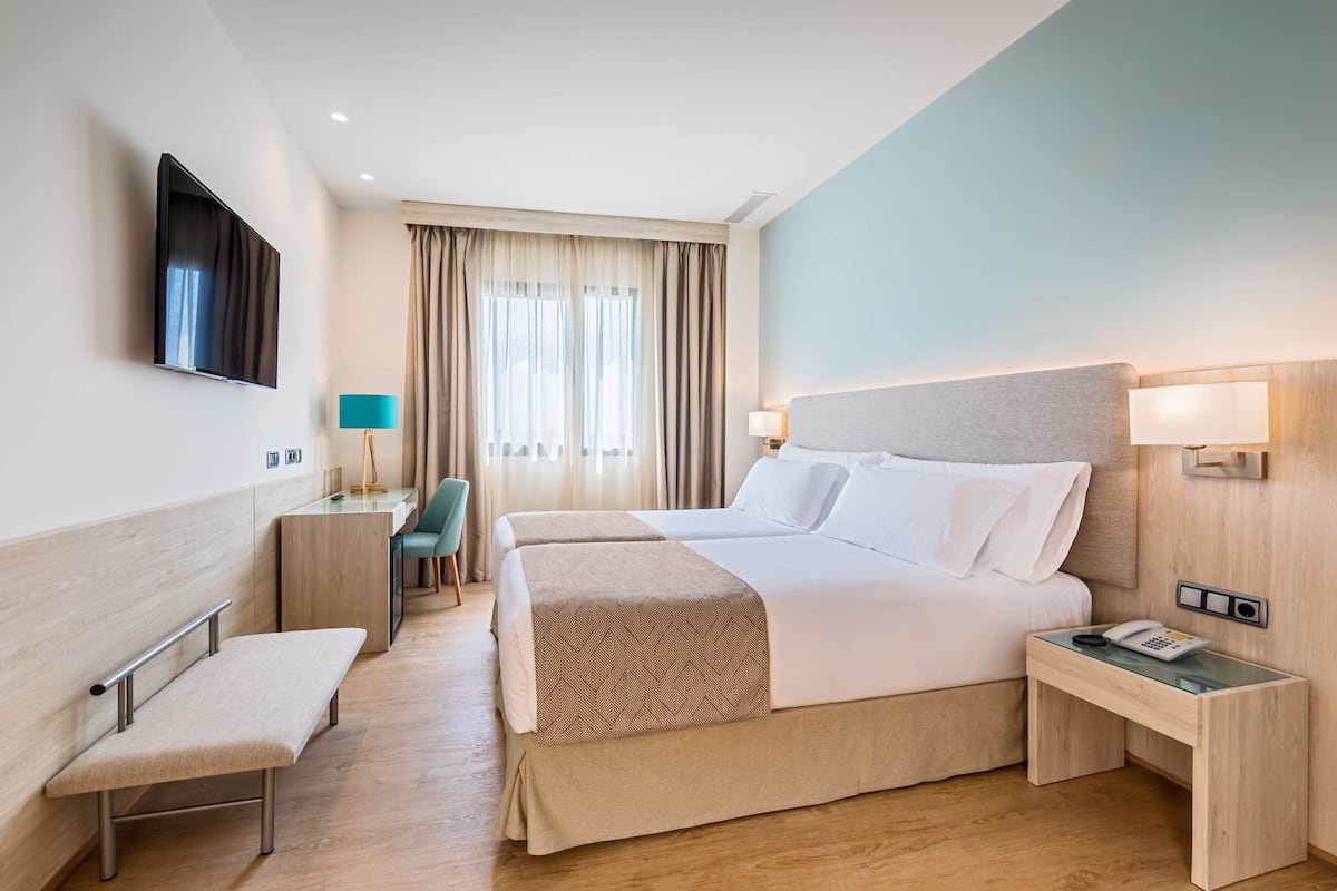 Ein Hotelzimmer mit zwei Betten, einem Schreibtisch, einer Bank und einem an der Wand montierten Fernseher.