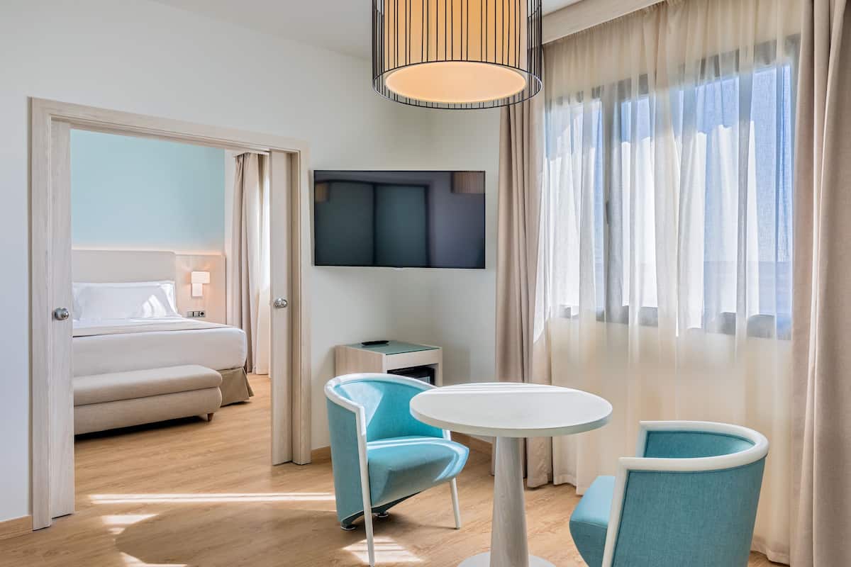 Ein modernes Hotelzimmer mit einem Bett, einem an der Wand montierten Fernseher, einem kleinen Tisch mit Stühlen und einem großen Fenster mit Vorhängen.