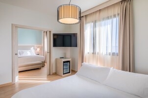 A hotel room with a large bed, a minibar, a flat-screen TV mounted on the wall, and a large window with curtains.