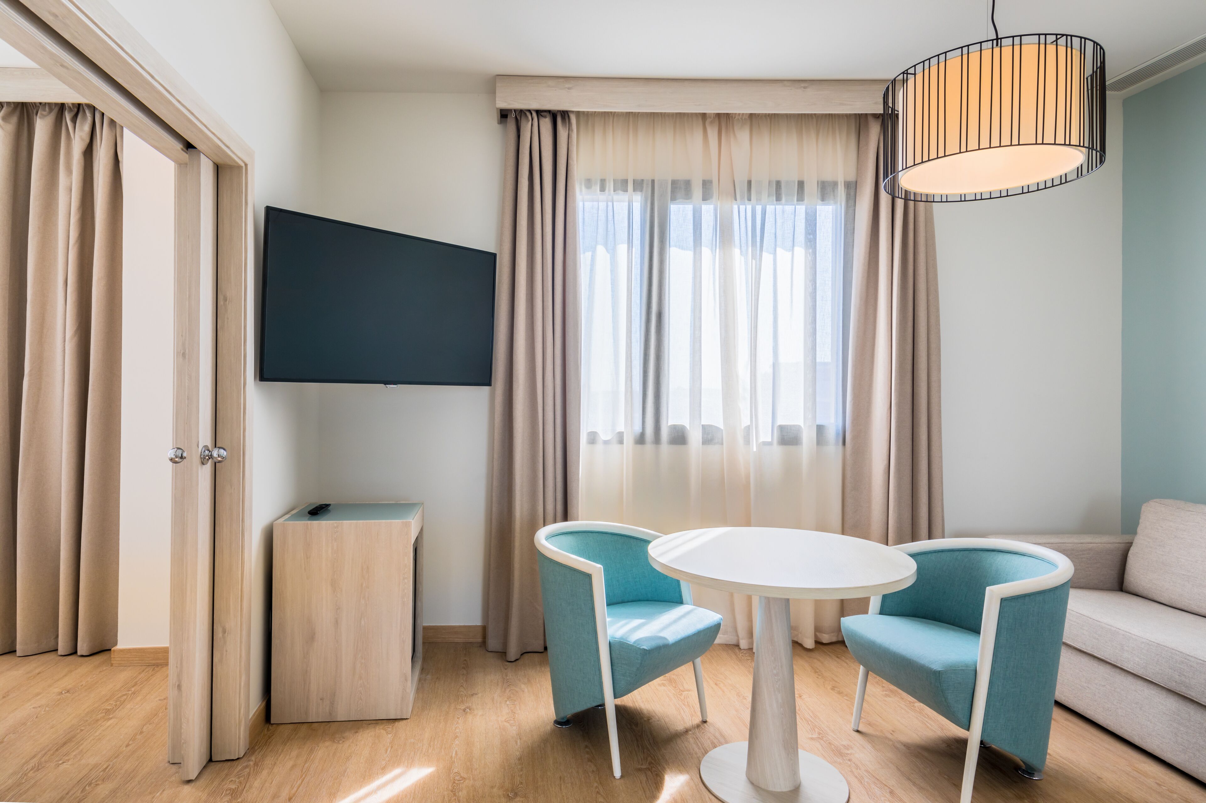 Habitación de hotel moderna con piso de madera, una mesa redonda blanca, dos sillones azul verdosos, un sofá color beige, un televisor montado en la pared y una lámpara en el techo.