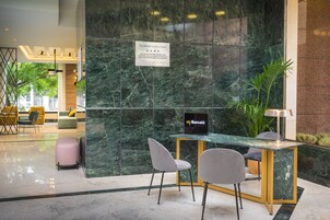 A modern hotel lobby with a marble reception desk, two grey chairs, a pink suitcase, and a sign indicating the hotel name.