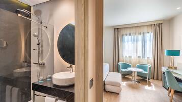 Un baño moderno con ducha envuelta en vidrio, un lavabo redondo y un espejo grande. Junto al baño, hay una zona de estar con dos sillas, una mesita pequeña, un ventanal con cortinas y una lámpara sobre una mesita auxiliar.