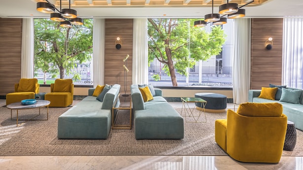 Een moderne woonkamer met een hoekbank, gele fauteuils en een salontafel. Grote ramen bieden uitzicht op bomen en een gebouw.