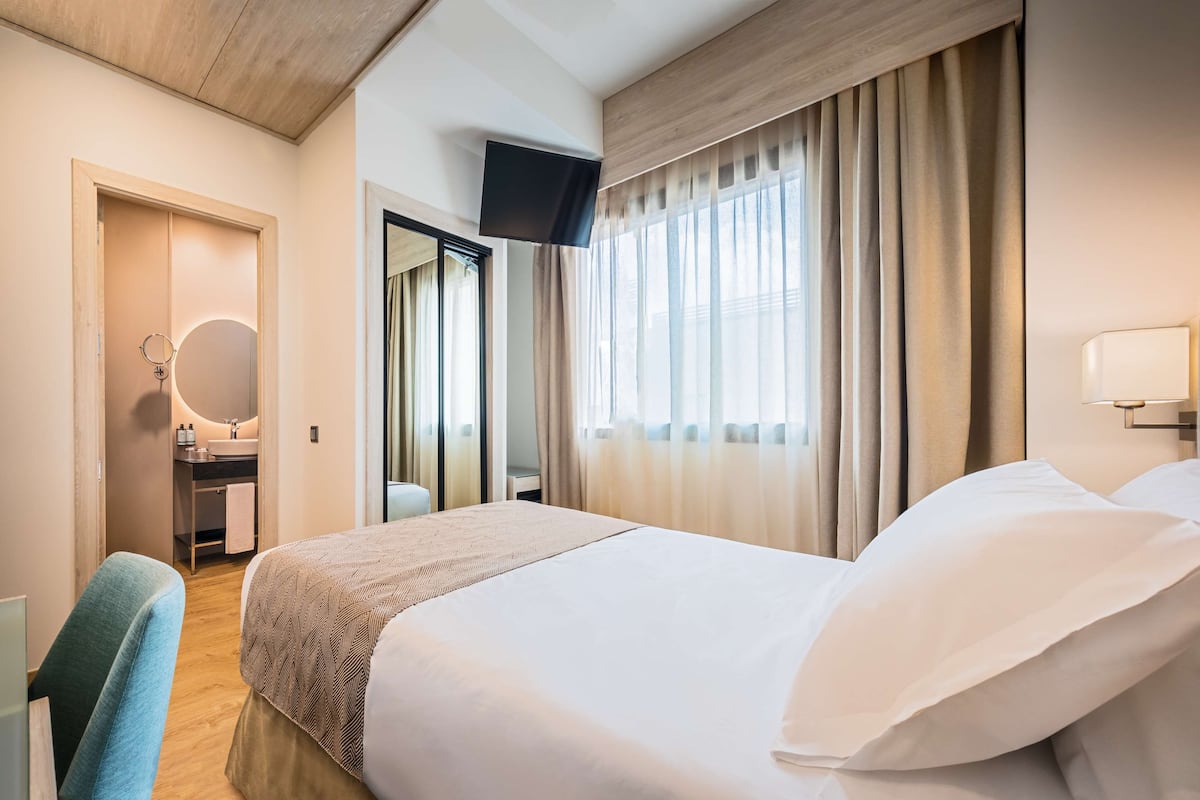 Ein Hotelzimmer mit einem Bett, einem Schreibtisch mit Stuhl, einem Spiegel, einem an der Wand befestigten Fernseher und einem Fenster mit Vorhängen.