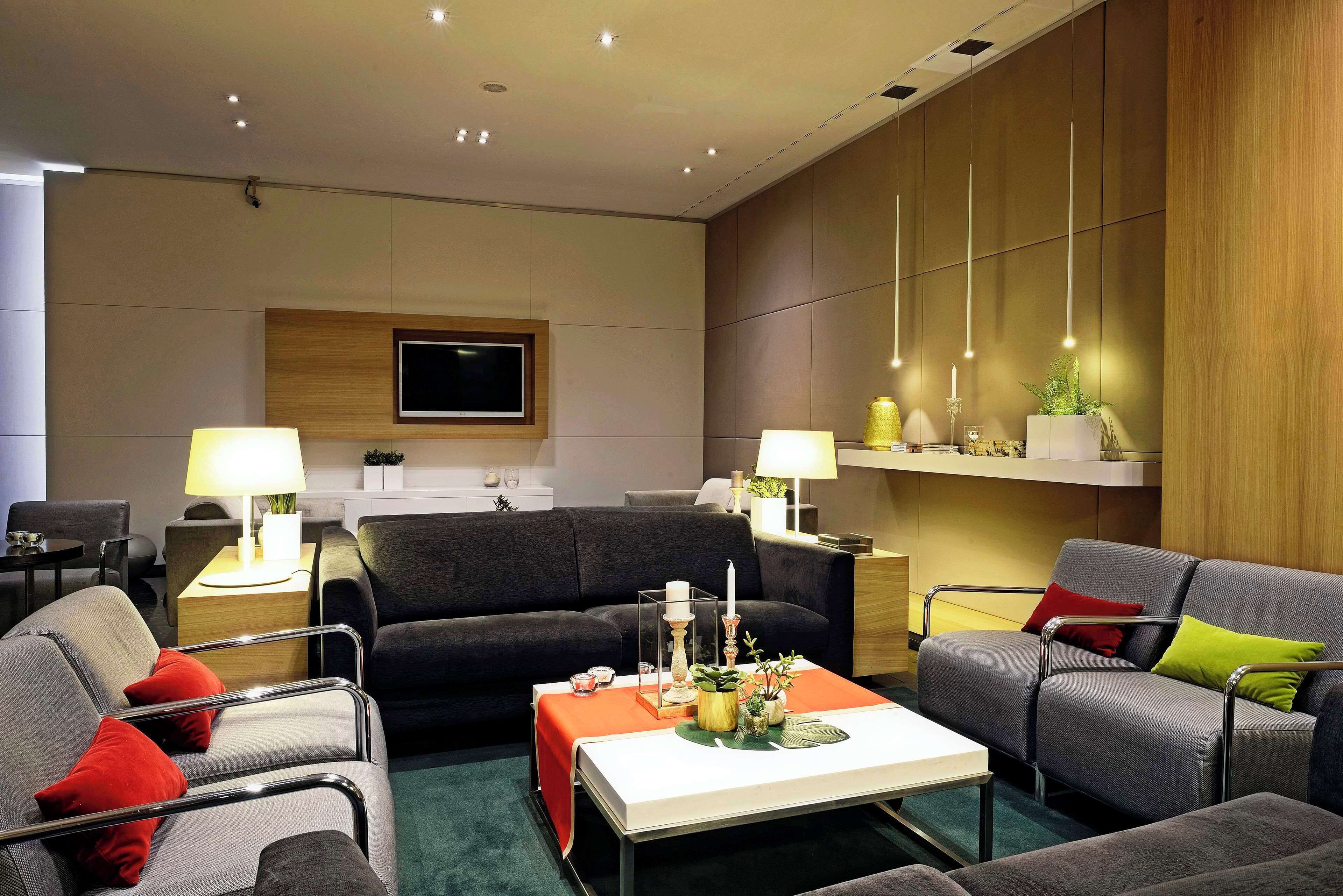 Ein modernes Wohnzimmer mit einem grauen Sofa, einem Couchtisch und zwei Sesseln. Auf den Nachttischen stehen zwei Lampen, an der Wand ist ein Flachbildfernseher und zur Rechten eine Holzverkleidung.