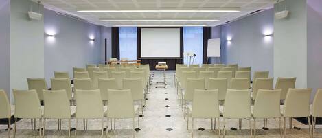 Un salón de conferencias con filas de sillas orientadas hacia una pantalla de proyección, una pizarra y una mesa con una computadora portátil.