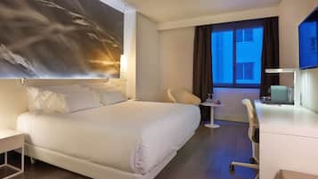 Habitación de hotel con una cama grande, un escritorio, una silla y vistas a la ciudad desde la ventana.