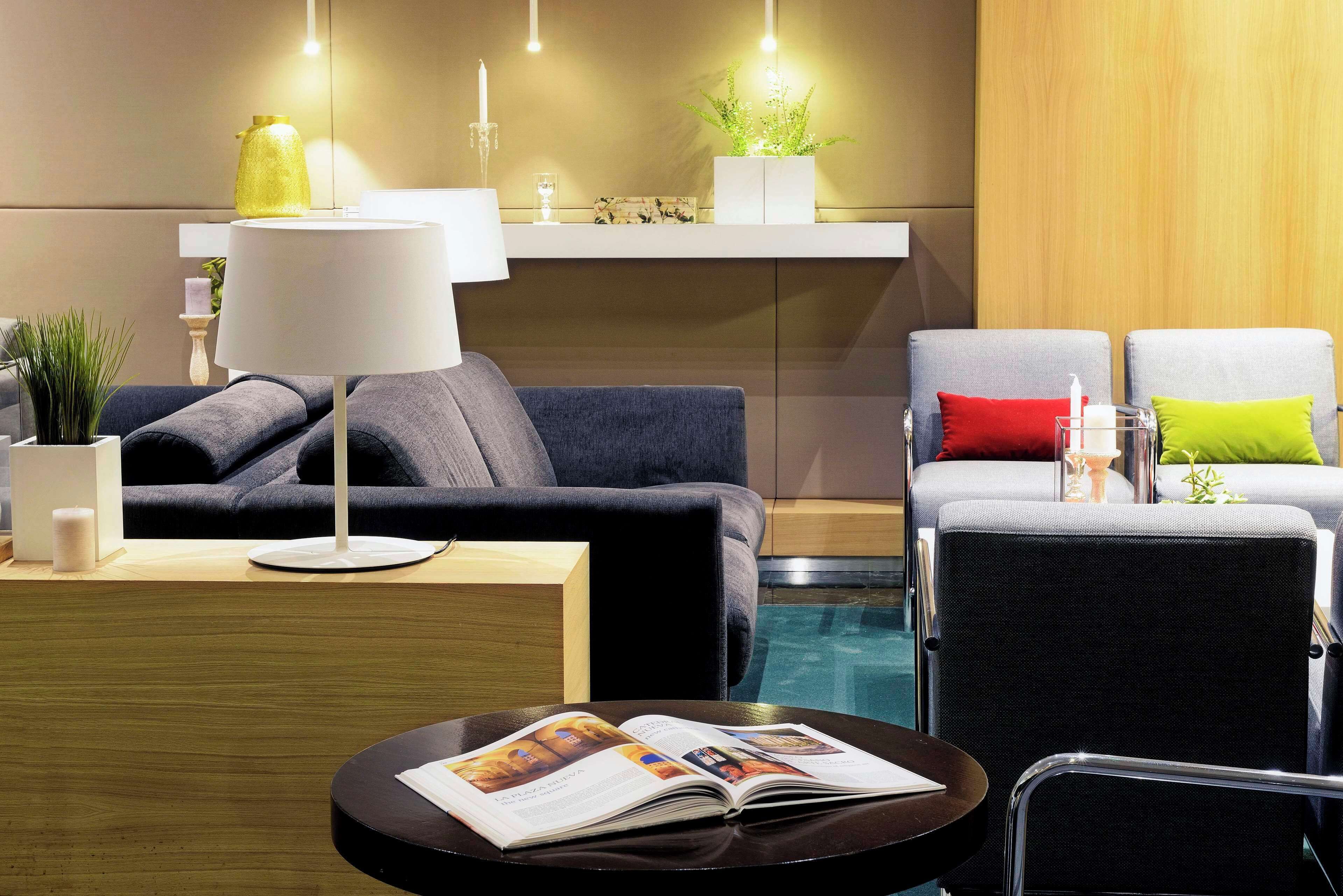 A modern hotel room with a dark grey sofa, a round black table with a magazine, a white lamp, and a small plant on a wooden shelf.