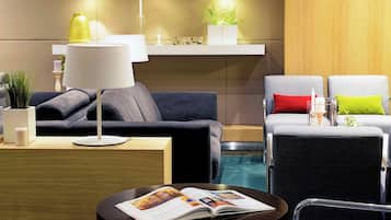 A modern hotel room with a dark grey sofa, a round black table with a magazine, a white lamp, and a small plant on a wooden shelf.