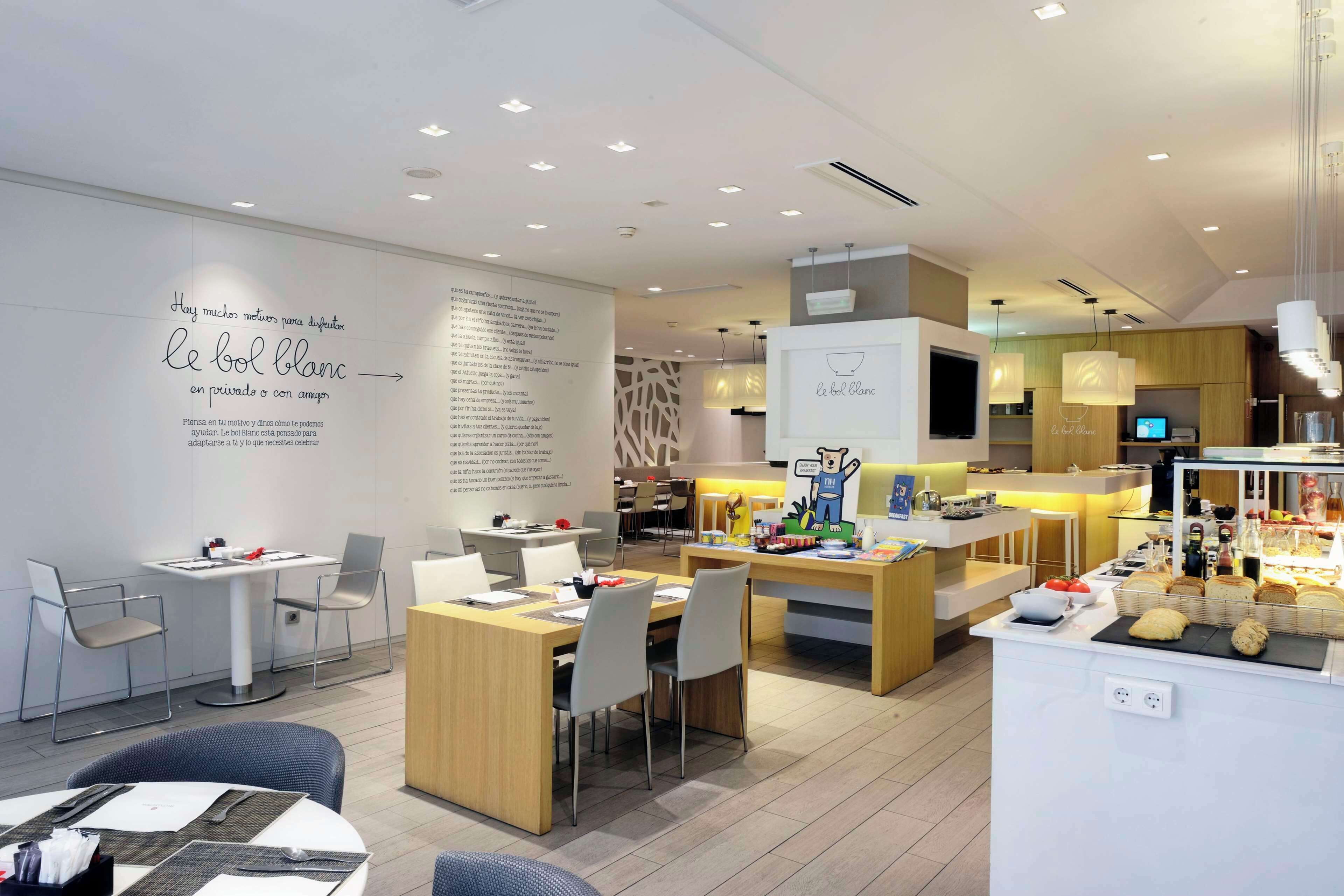 Una cafetería moderna con un diseño minimalista, que presenta una barra central con una pantalla de menú, zonas de estar con mesas y sillas, y una pared con texto y gráficos decorativos.
