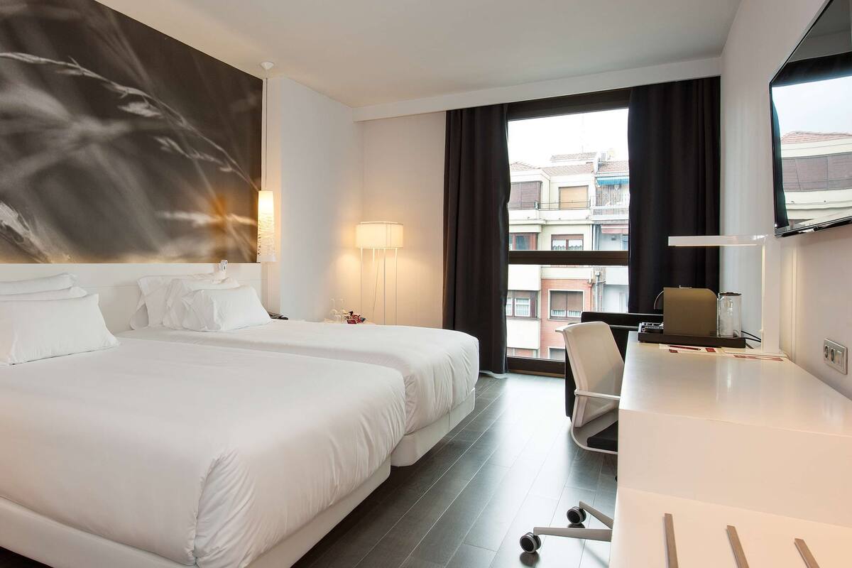 Une chambre d’hôtel avec deux lits, un bureau, une chaise, une grande fenêtre et une œuvre d’art accrochée au mur.