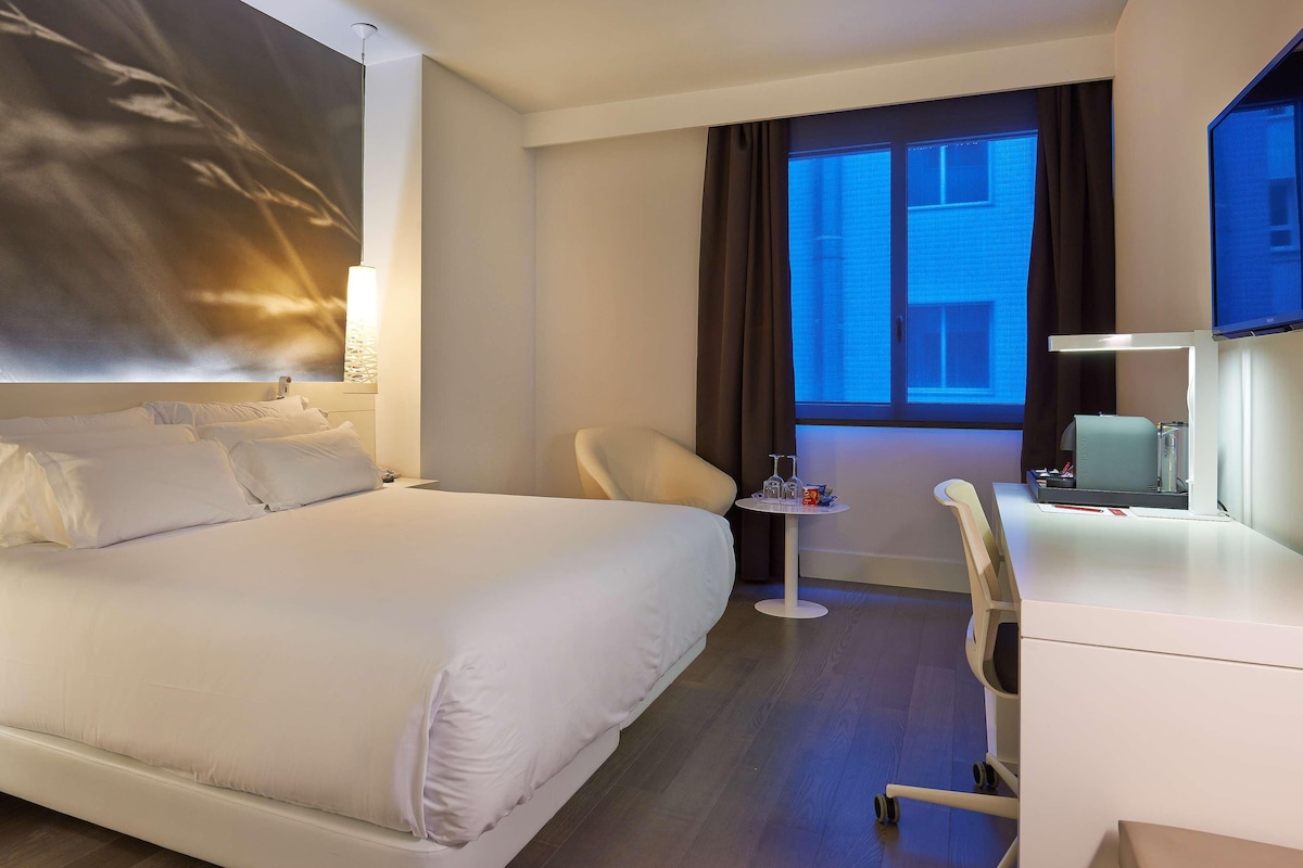 A modern hotel room with a large bed, a desk, a chair, and a television.