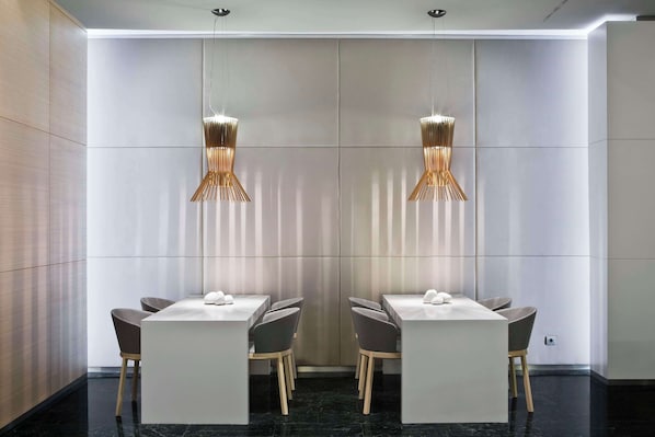 Una sala de conferencias moderna con una distribución simétrica, que presenta una mesa central con sillas, dos lámparas colgantes y un amplio panel en la pared.