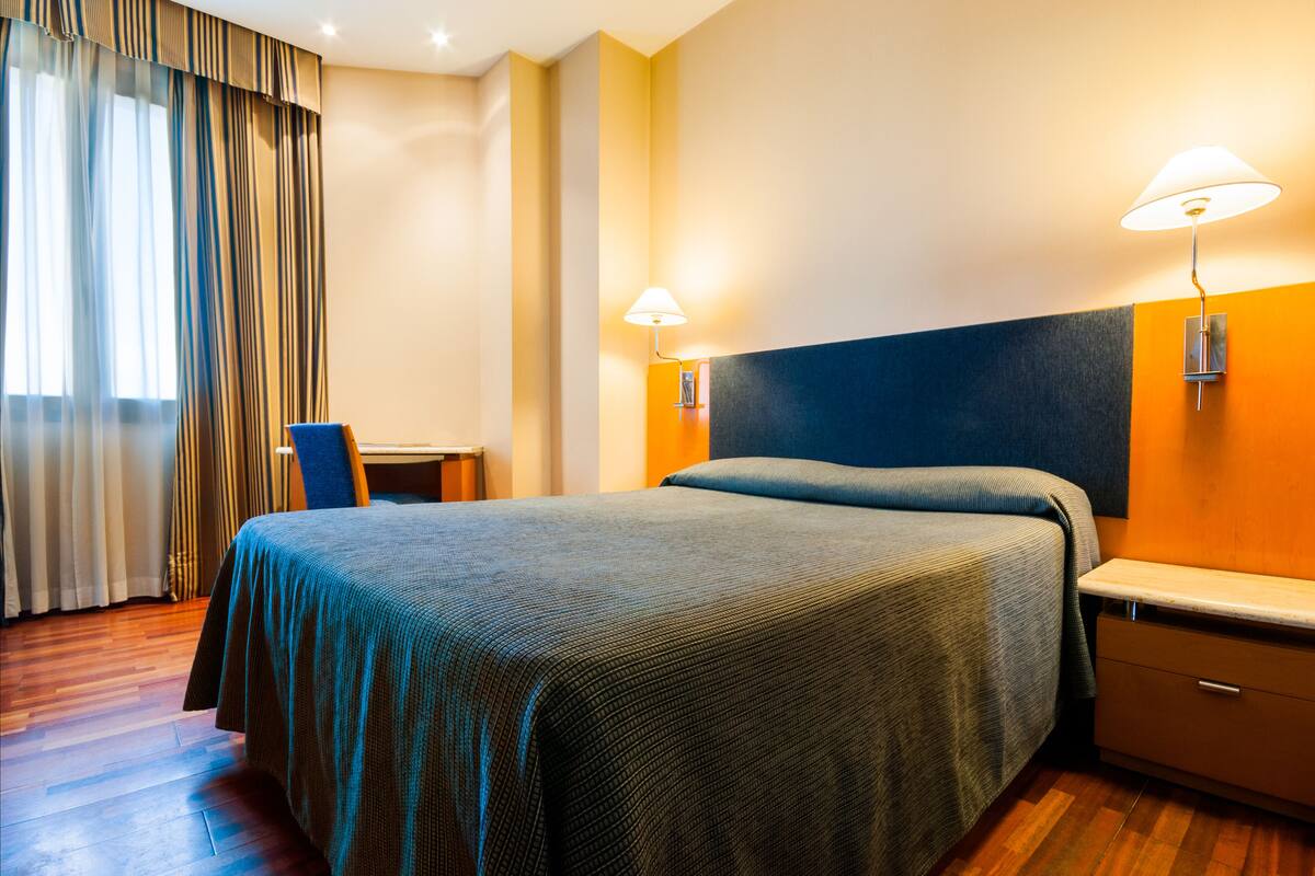 A hotel room with a bed, nightstand, lamp, chair, and curtains.