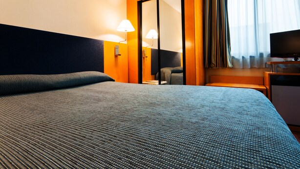 A hotel room with a large bed, a desk with a television, a chair, and a mirror. - Hotel Villacarlos (Valencia)