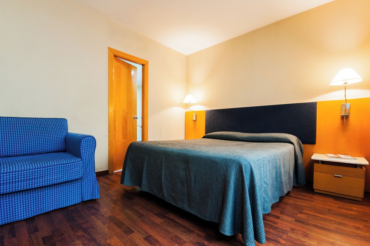 A hotel room with a bed, a blue armchair, a wooden nightstand, and two wall-mounted lamps.