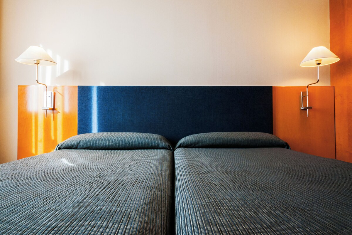 A hotel room with two beds, a blue headboard, and wall-mounted lamps.