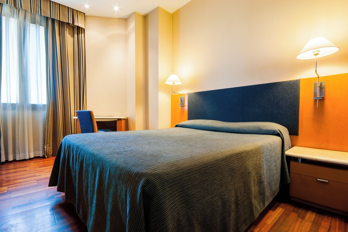 A hotel room with a bed, a desk, and two wall-mounted lamps.