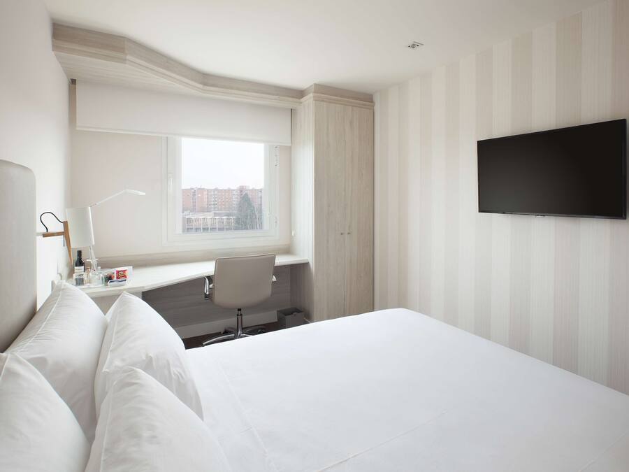 A hotel room with a large bed, a flat-screen TV mounted on the wall, a window with a view of buildings, a desk with a lamp, and a chair.
