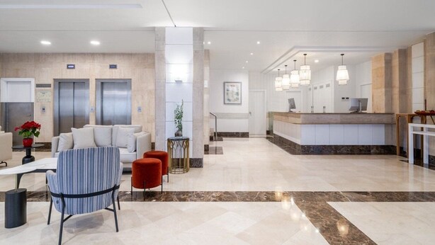 Un moderno vestíbulo de hotel con mostrador de recepción, zona de estar, ascensores y suelo de mármol.