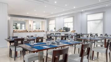 Un vaste espace dédié à la salle à manger, avec de nombreuses tables et chaises, une cuisine à l’arrière-plan et de grandes fenêtres qui laissent entrer la lumière naturelle.