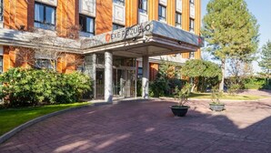 La entrada de un hotel con vestíbulo cubierto, un cartel con el nombre y un área ajardinada con plantas en macetas.