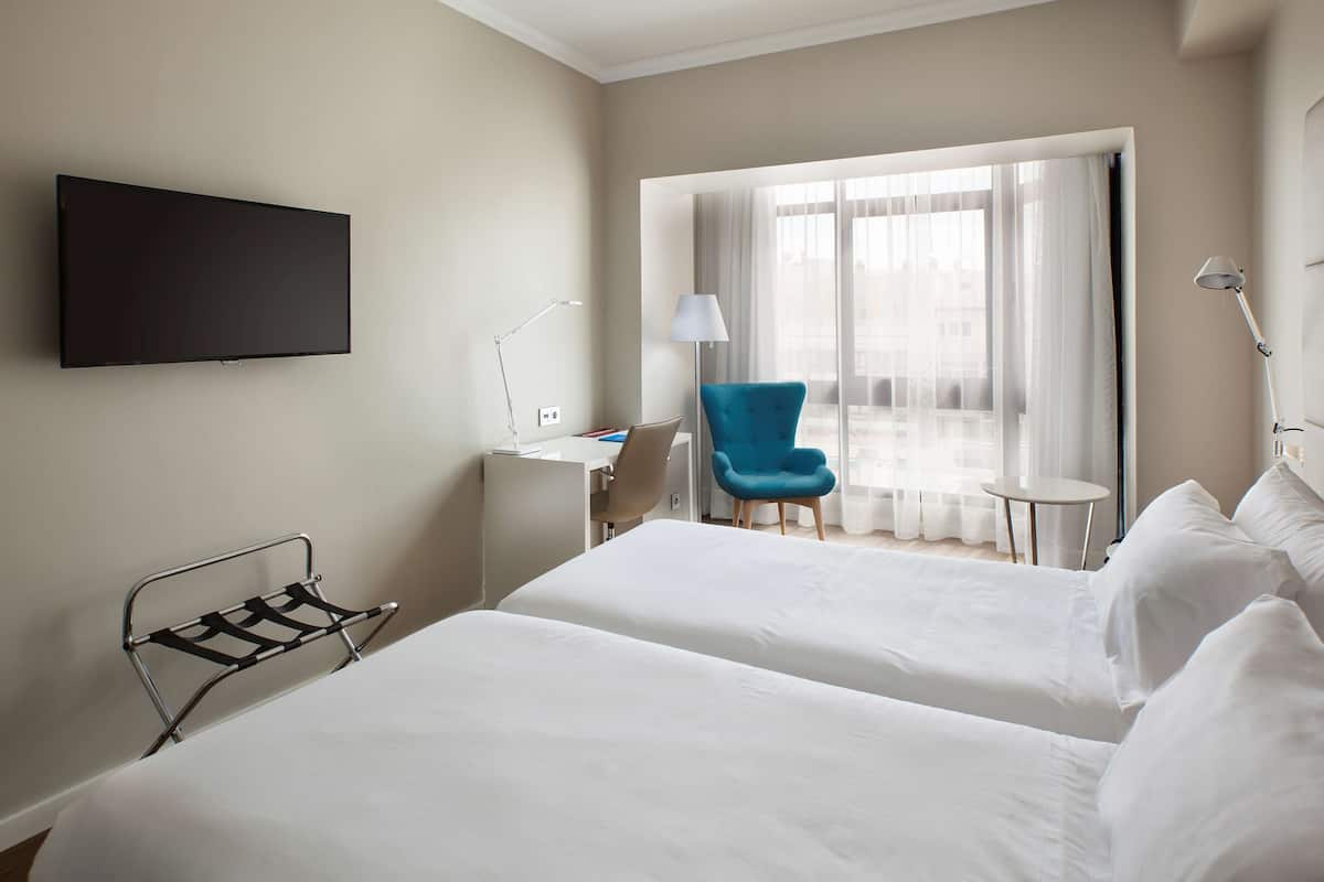 Une chambre d’hôtel avec deux lits, un bureau, une chaise, une télévision et une fenêtre avec des rideaux.