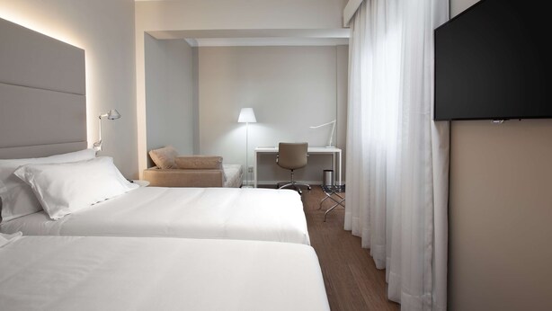 A hotel room with two beds, a desk, a chair, a lamp, and a TV.