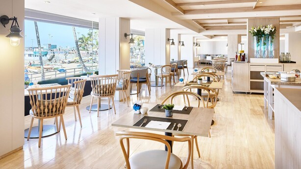 A modern restaurant with wooden furniture, large windows offering a view of a pool and palm trees, and a kitchen area visible in the background. - NH Las Palmas Playa las Canteras (Las Palmas de Gran Canaria)