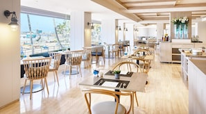 Un restaurante moderno con muebles de madera, grandes ventanales que dan a una piscina y palmeras, y una cocina visible al fondo.