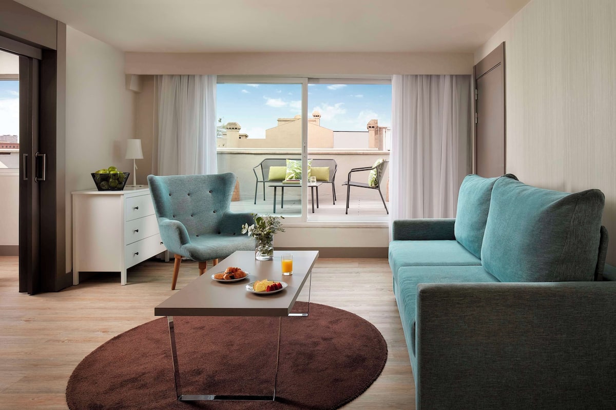Una habitación de hotel moderna con sofá, sillón, mesa de comedor y balcón con vistas a edificios.