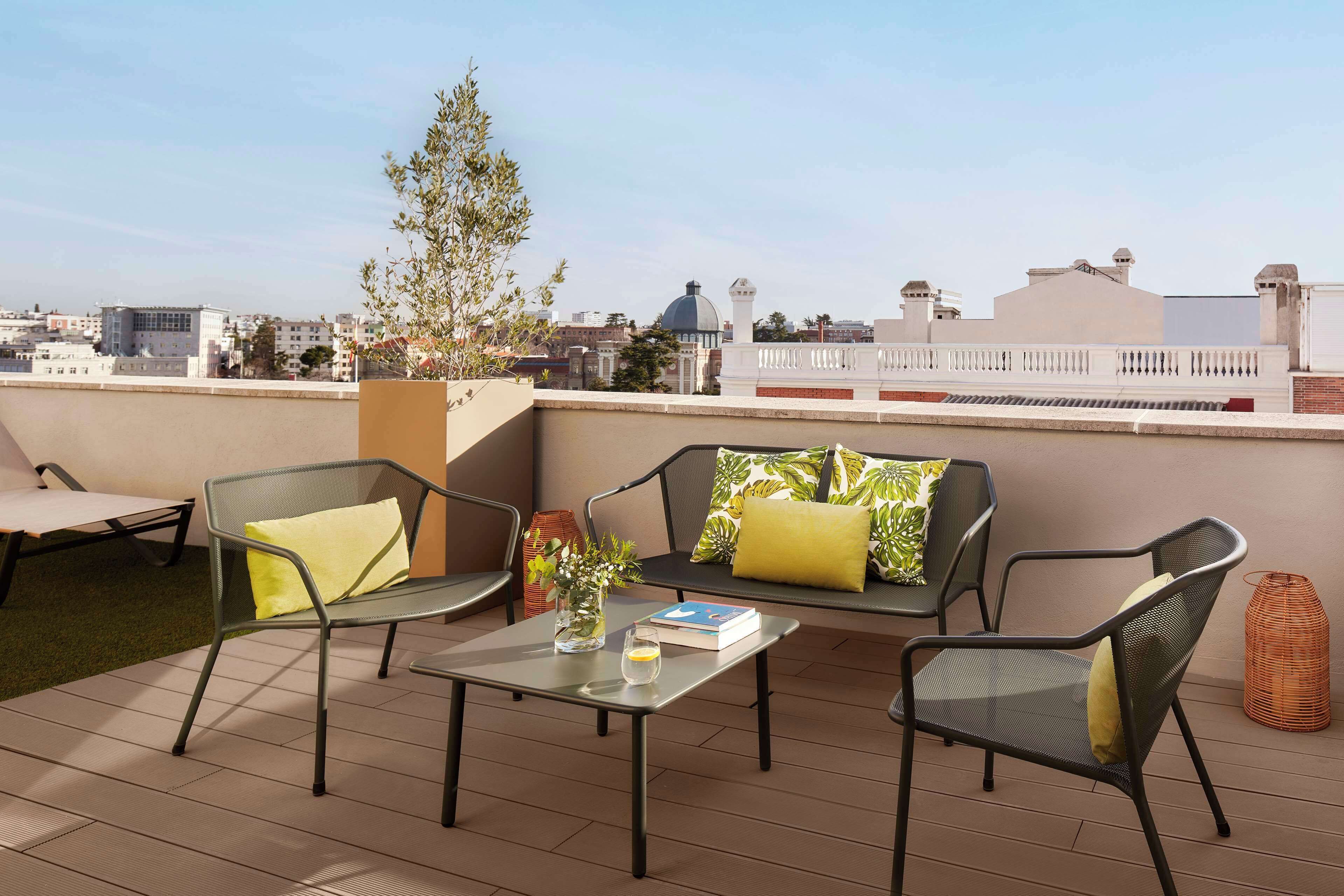 A rooftop terrace with outdoor furniture, a small table with a book and a vase, and a view of the cityscape.