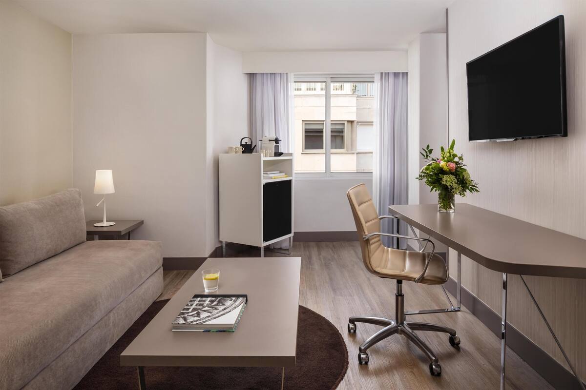 Habitación de hotel con sofá, una mesa de centro con una revista, un escritorio con una silla, televisor montado en la pared y una ventana con cortinas.