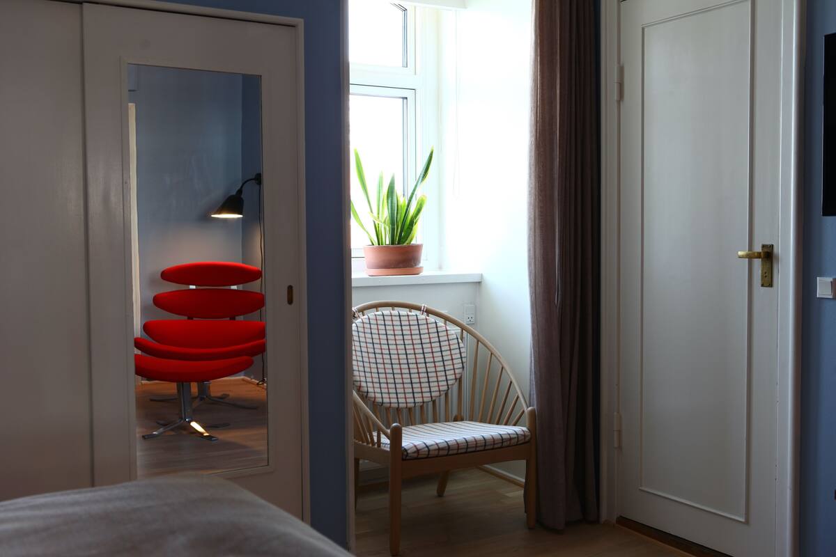A room with a wooden chair, a red chair, a potted plant, and a window with curtains.
