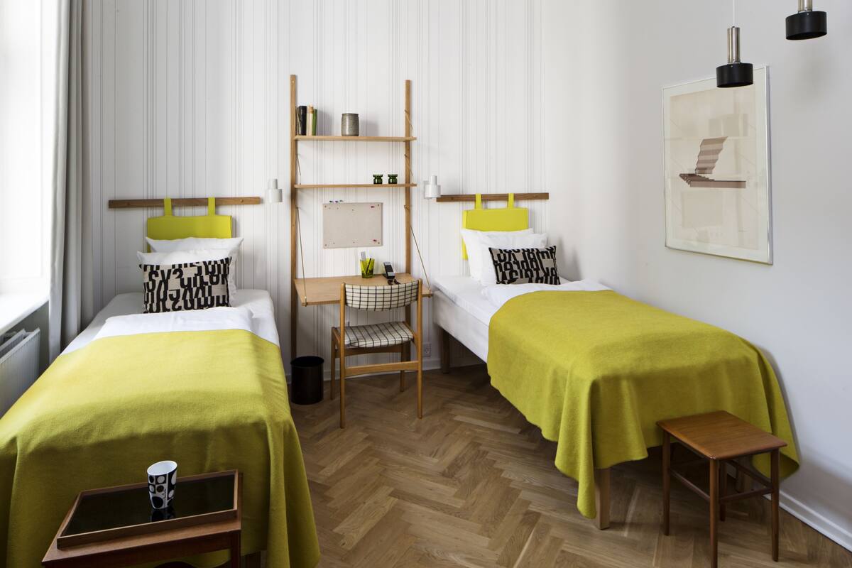A room with two beds, a small desk, a chair, and a shelf with items.