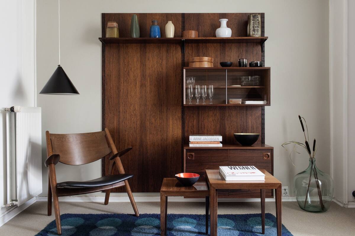 A room with a wooden chair, a small table with a bowl and books, a bookshelf with decorative items, and a glass vase with plants.