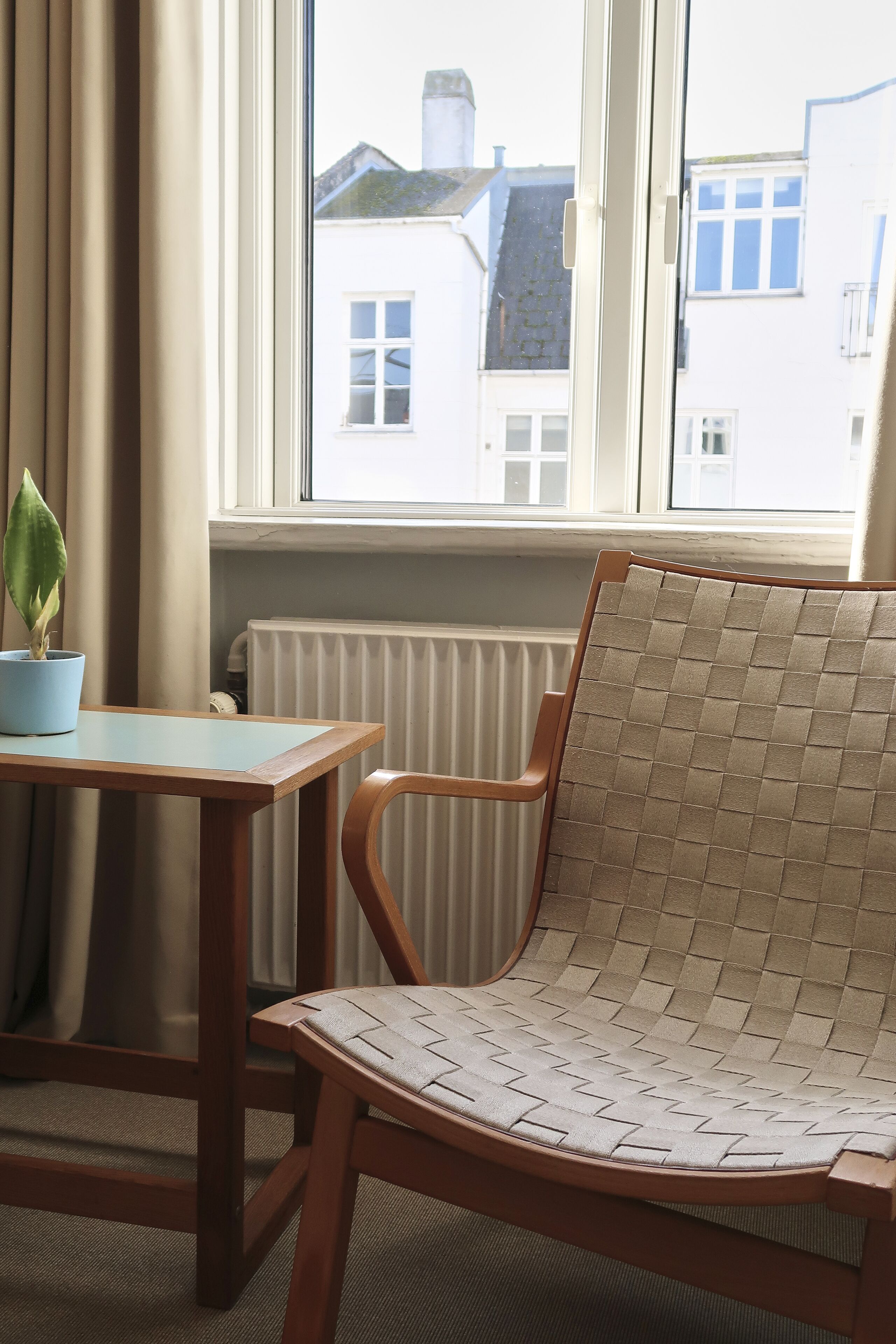 Et værelse med en træstol, et lille bord, en potteplante og et vindue med udsigt til bygninger.