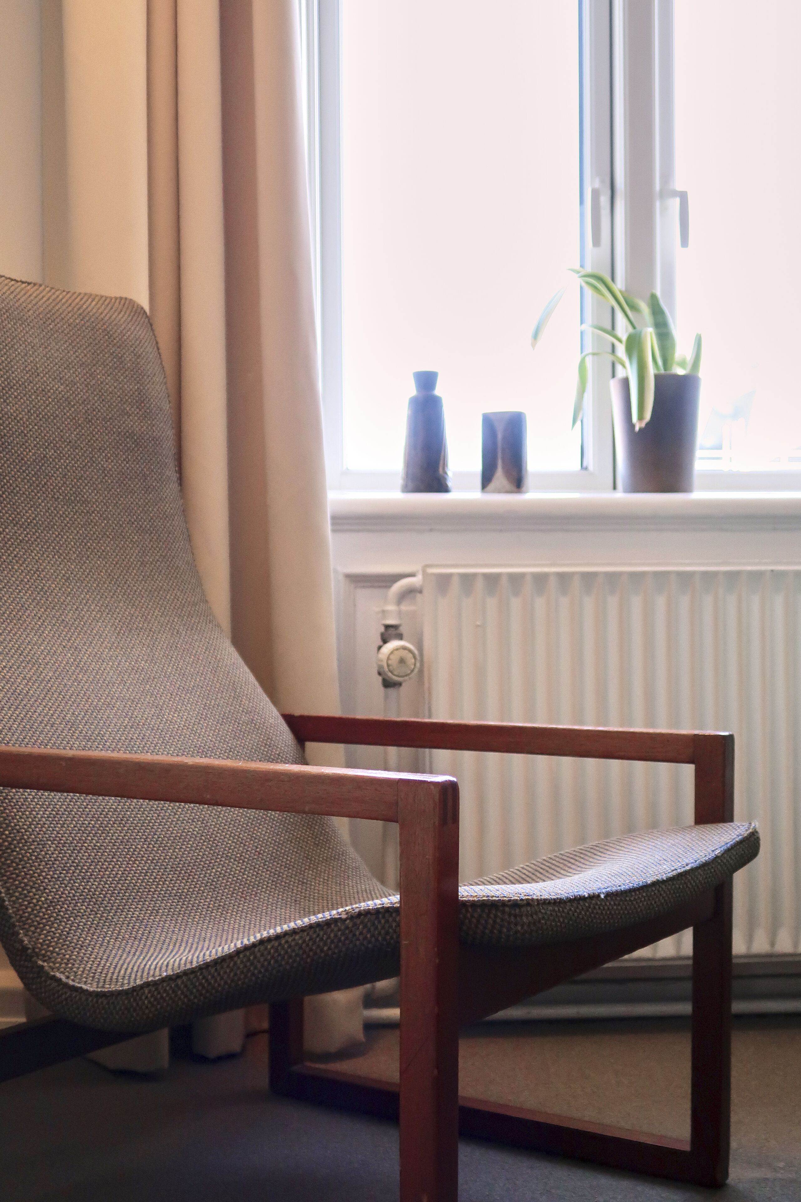 Ein Stuhl mit gewobener Sitzfläche und Lehne, vor einem Fenster mit Vorhängen, einem Heizkörper und einem Blumentopf auf der Fensterbank.