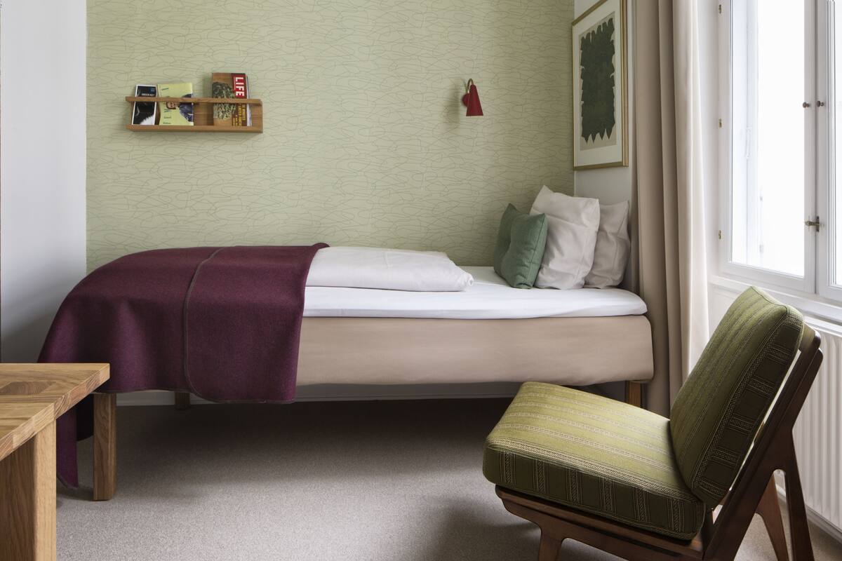 A neatly arranged bedroom with a bed, a chair, a small table, and a shelf with books.