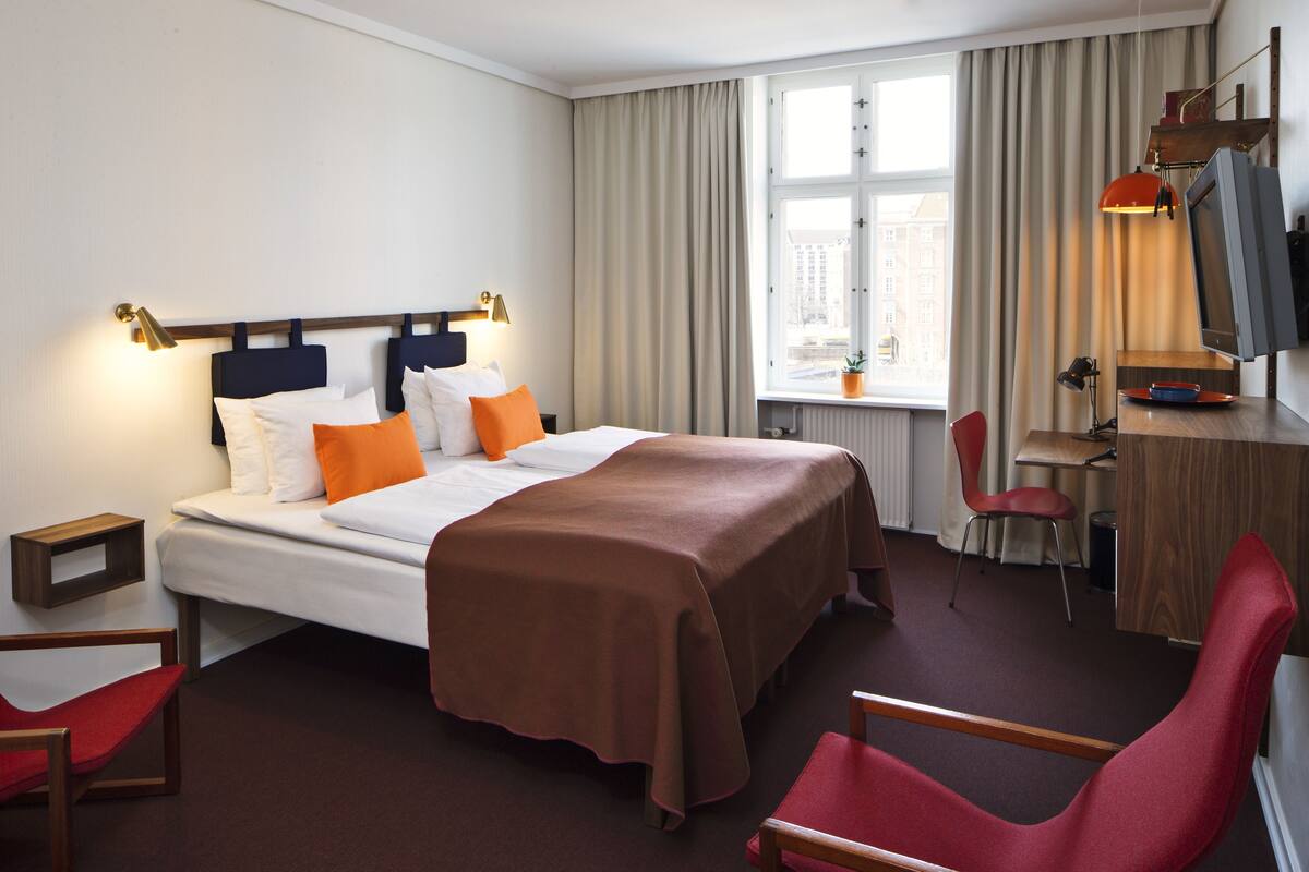 A hotel room with a bed, a desk, a chair, a TV, and a window with curtains.