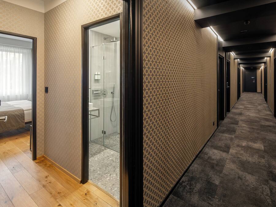 A hotel corridor with a patterned wall, a glass door leading to a bathroom, and a wooden floor.