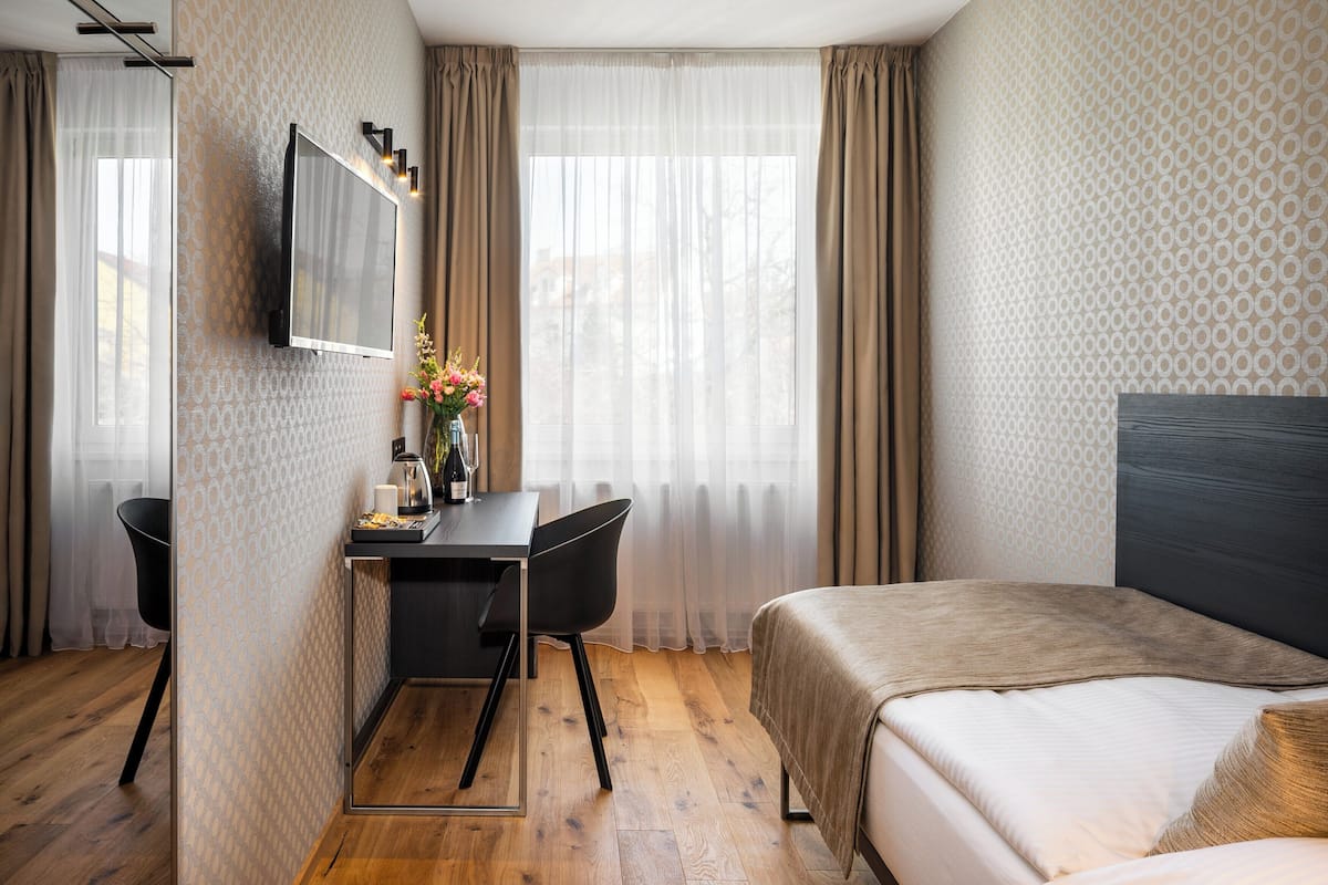 Habitación de hotel con cama, escritorio y silla, televisión montada en la pared y ventana con cortinas.