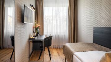 A hotel room with a bed, a desk with a chair, a TV mounted on the wall, and a window with curtains.