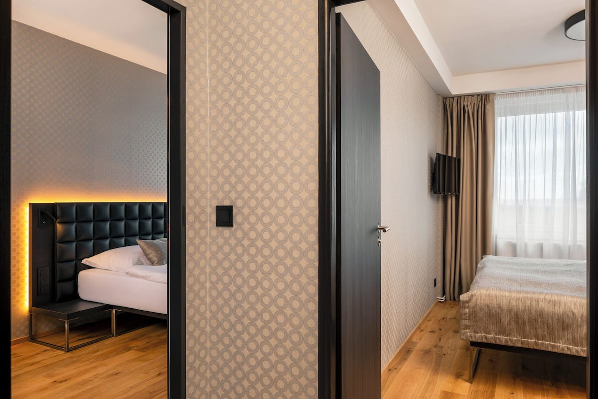 Una habitación de hotel moderna con una cama, una mesita de noche, un televisor montado en la pared y una puerta que da a otra habitación.