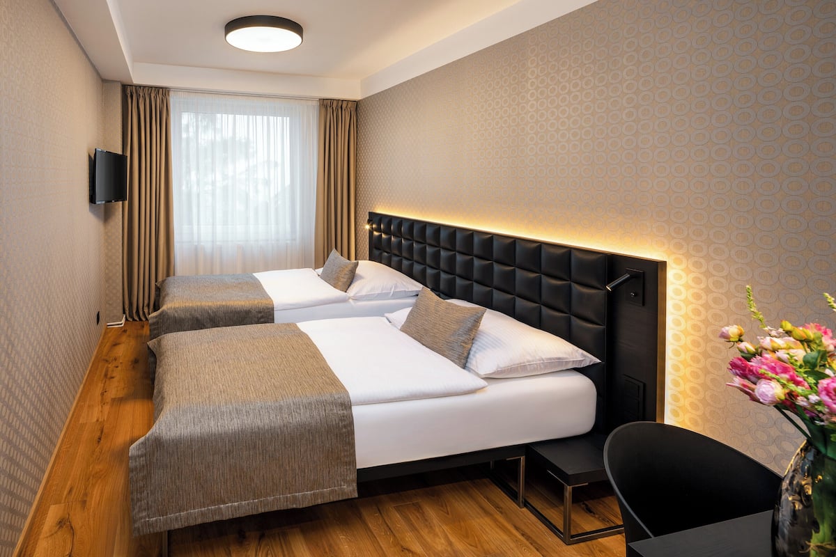 Habitación de hotel con dos camas, un televisor de pantalla plana montado en la pared, piso de madera y un florero con flores sobre la mesita de noche.