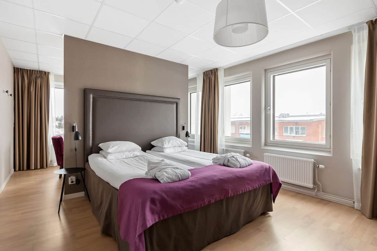 A modern hotel room with a large bed, a bedside table, a lamp, and a view of the city through the window.